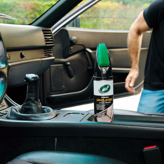 Car interior shot: Turtle Wax upholstery cleaner with brush applicator sits near a black leather gear stick and dark dashboard.