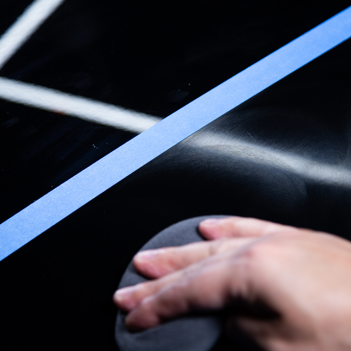 Dark paint correction. Auto detailing shows before and after paintwork by a hand holding a detailing pad next to masking tape.
