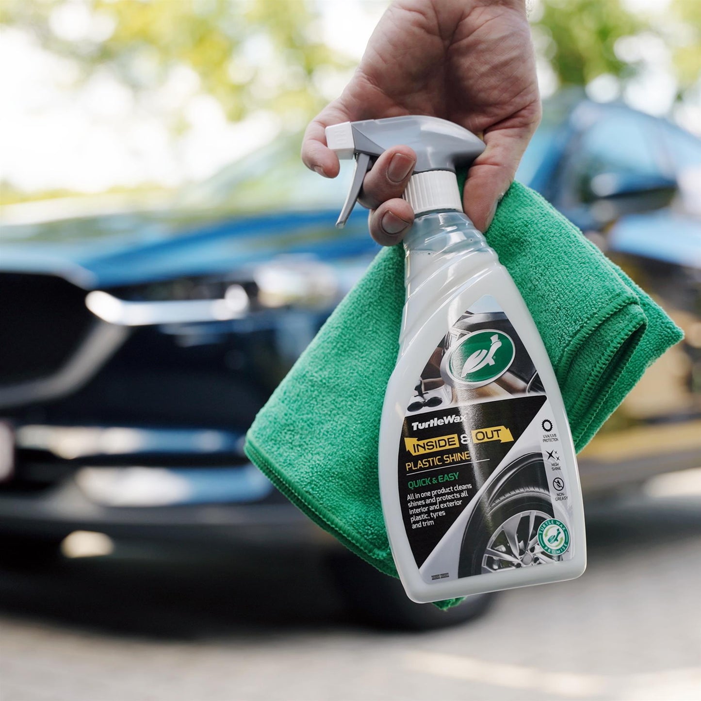 Hand holding Turtle Wax cleaner and a cloth in front of a car. Car care for interiors, exteriors, plastic, tyres and trim.