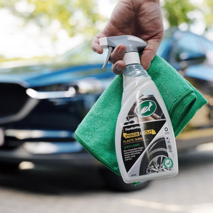 Hand holding Turtle Wax cleaner and a cloth in front of a car. Car care for interiors, exteriors, plastic, tyres and trim.