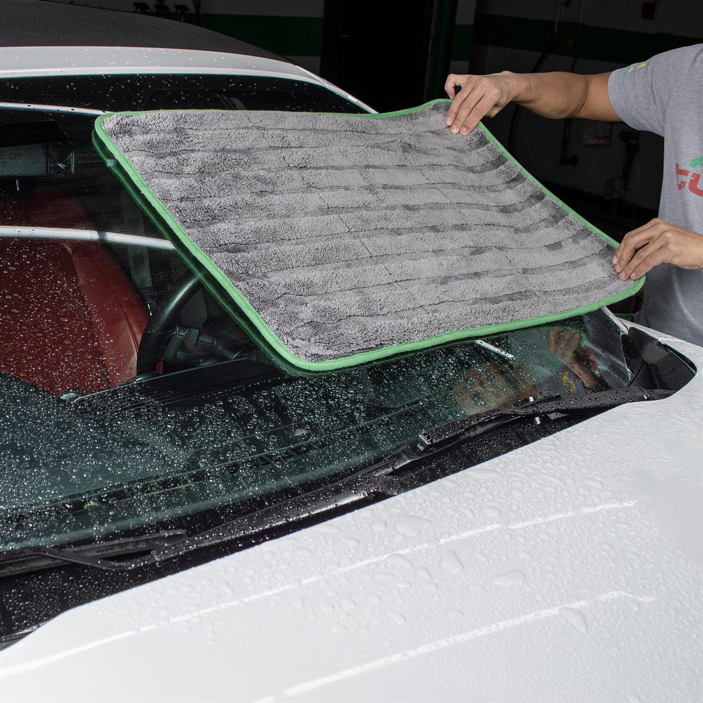Car detailer uses grey Turtle Wax microfibre drying towel with green trim on windscreen of wet white car.