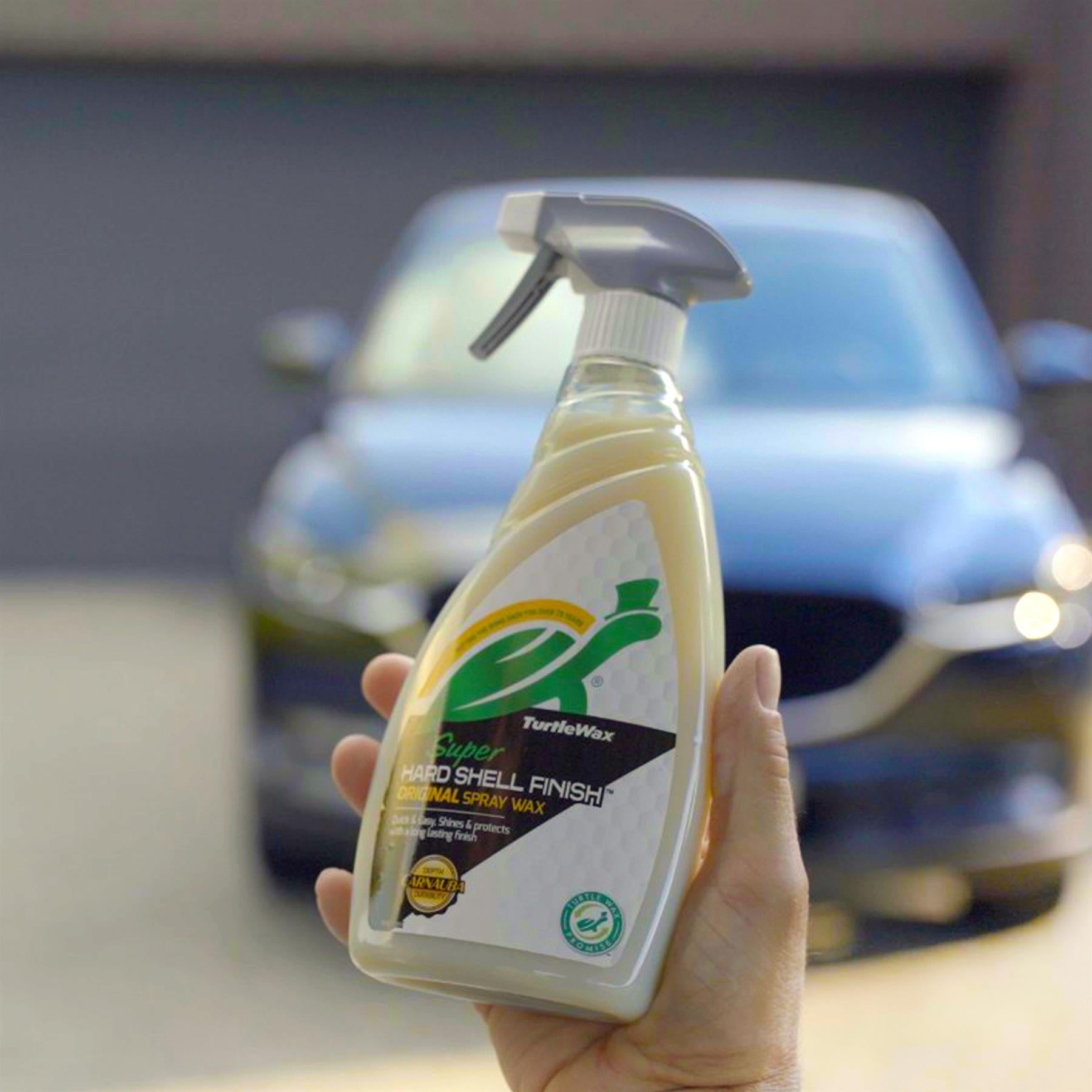 Hand holds Turtle Wax "Super Hard Shell Finish" spray. Car polish with Carnauba wax for car detailing shown in front of a blurry vehicle.
