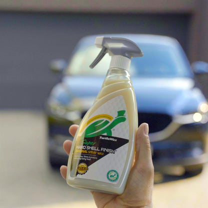 Hand holds Turtle Wax "Super Hard Shell Finish" spray. Car polish with Carnauba wax for car detailing shown in front of a blurry vehicle.
