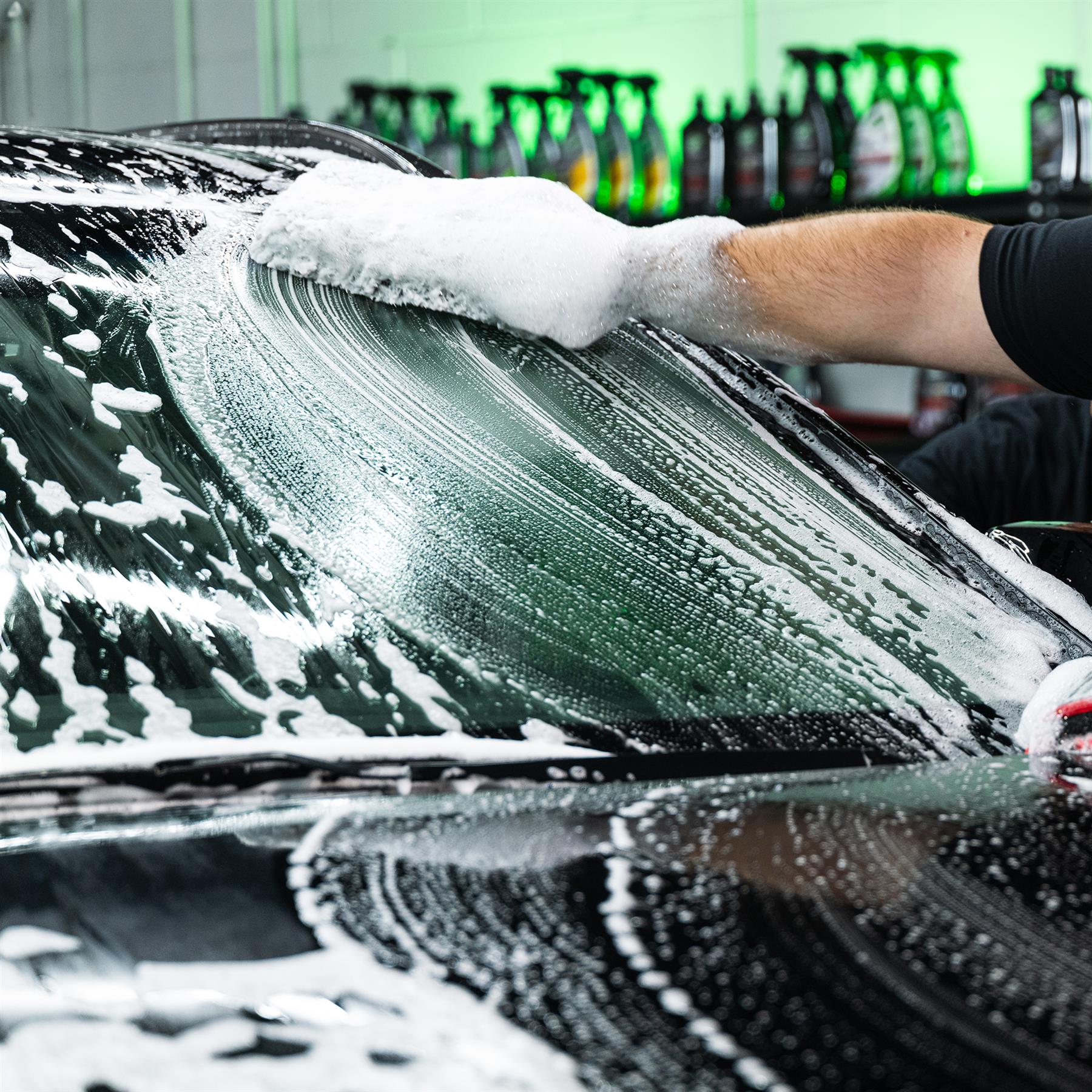 Car cleaning. A gloved hand washes a dark car's windscreen with soapy suds in a detailing shop.
