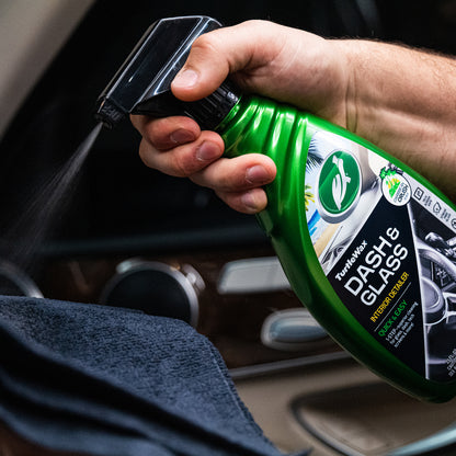 Hand sprays Turtle Wax dash & glass cleaner onto a cloth for car interior detailing. Focus on cleaning glass and dashboard surfaces.
