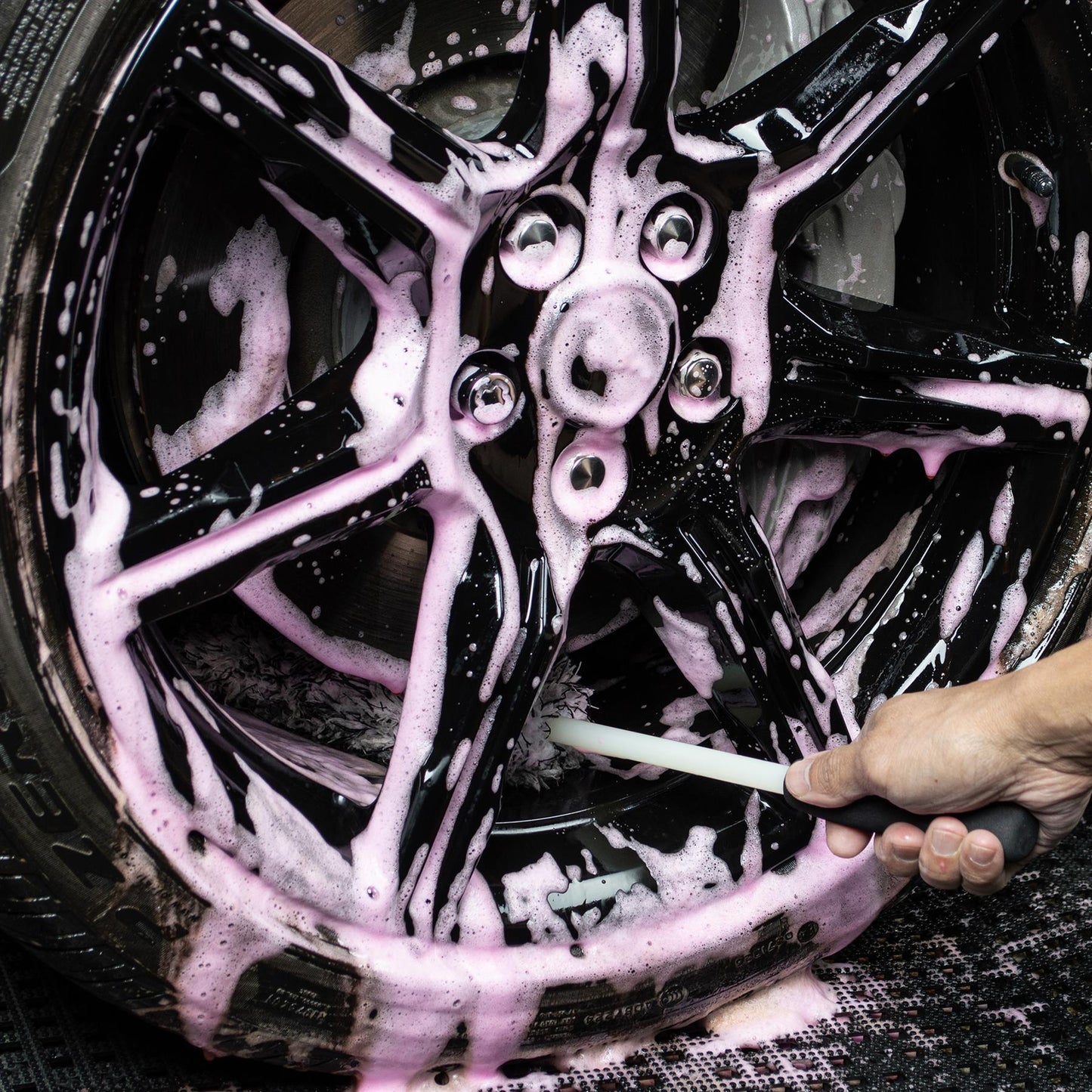 Close-up of black alloy wheel being cleaned with a brush and pink foamy Turtle Wax solution, detail shot.