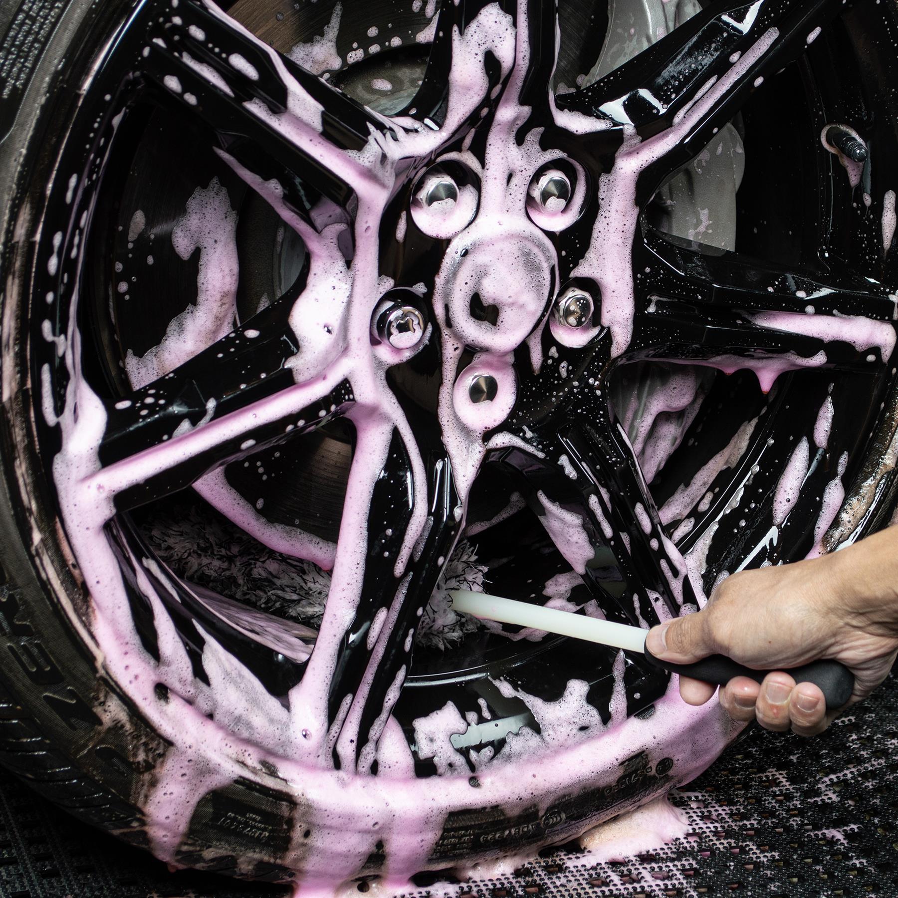 Close-up of black alloy wheel being cleaned with a brush and pink foamy Turtle Wax solution, detail shot.
