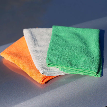 Three microfiber cloths in orange, gray, and green on a reflective surface with a dark background.
