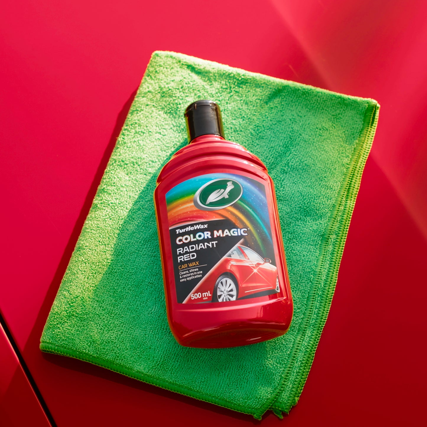 Turtle Wax Color Magic Radiant Red, a car wax, rests on a green microfibre cloth atop a red car panel, promising shine and restoration.
