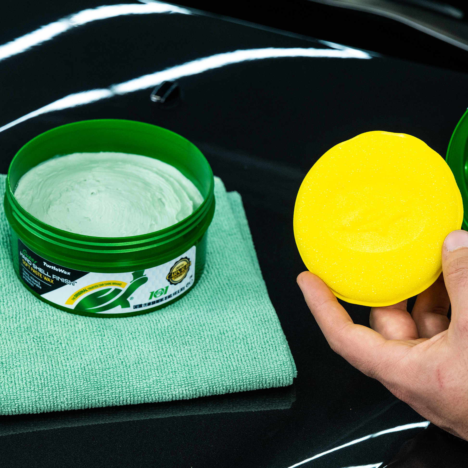 Turtle Wax Super Hard Shell paste on a black car bonnet. A yellow foam pad and green microfibre cloth are nearby. Car care detail.
