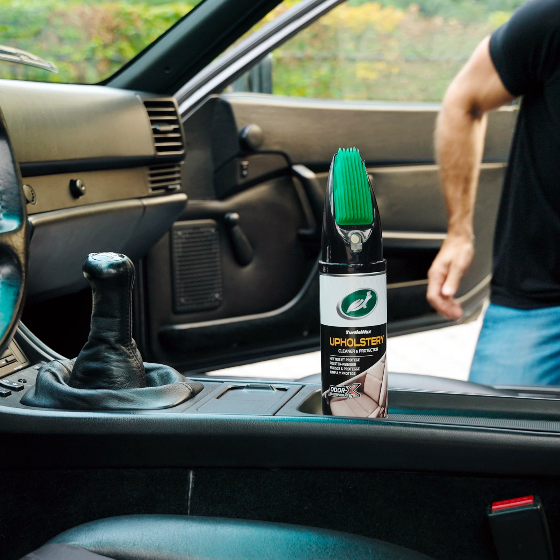 Car interior shot: Turtle Wax upholstery cleaner with brush applicator sits near a black leather gear stick and dark dashboard.