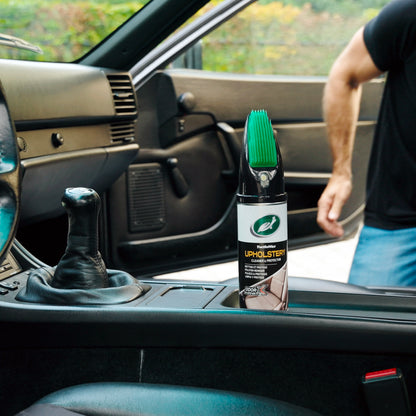 Car interior shot: Turtle Wax upholstery cleaner with brush applicator sits near a black leather gear stick and dark dashboard.