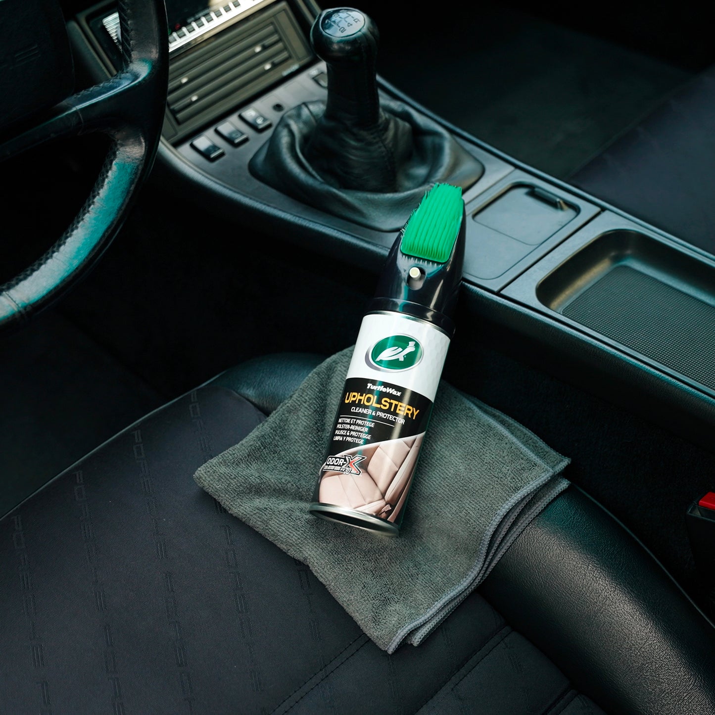 Turtle Wax upholstery cleaner with applicator brush sits on a cloth in a car interior by the gear stick and steering wheel.