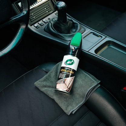 Turtle Wax upholstery cleaner with applicator brush sits on a cloth in a car interior by the gear stick and steering wheel.