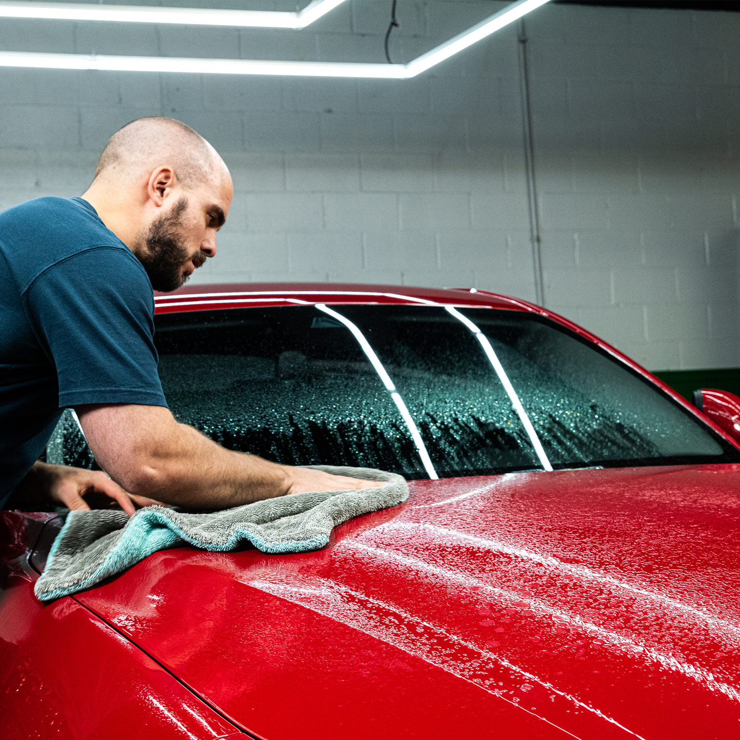 Red car detailing. A man wiping a wet, glossy red car with a grey microfibre cloth.