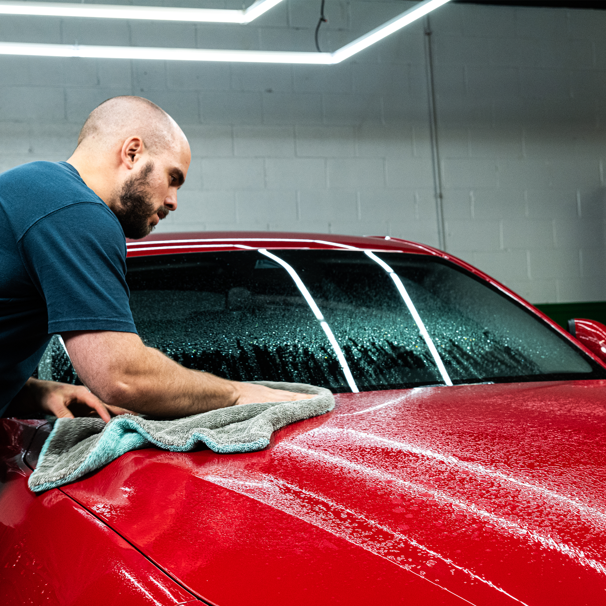 Red car detailing. A man wiping a wet, glossy red car with a grey microfibre cloth.