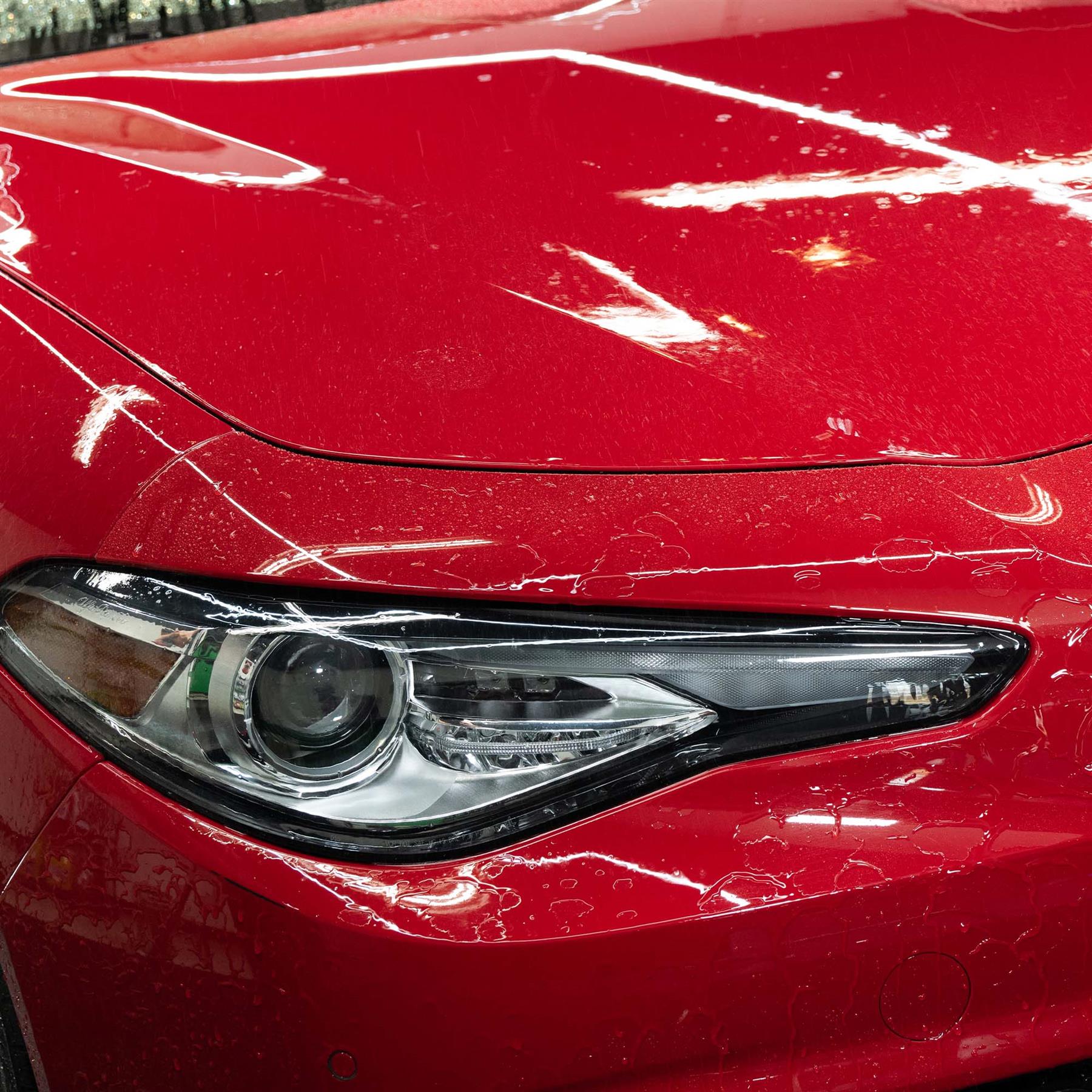 Red car detail. Water droplets show fresh wash sheen. Modern headlight visible.
