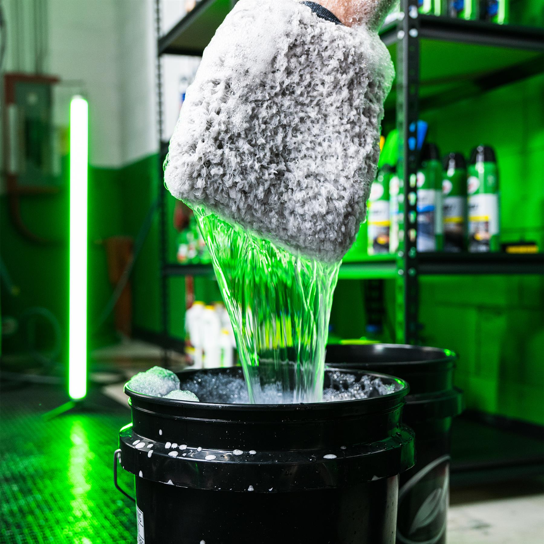 Soapy mitt above black buckets with car wash solution. Green liquid pours down. Cleaning accessories are on shelving.