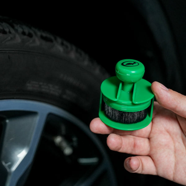 Turtle Wax applicator brush for tyre shine held in hand near a car wheel. The applicator is green with a circular brush.
