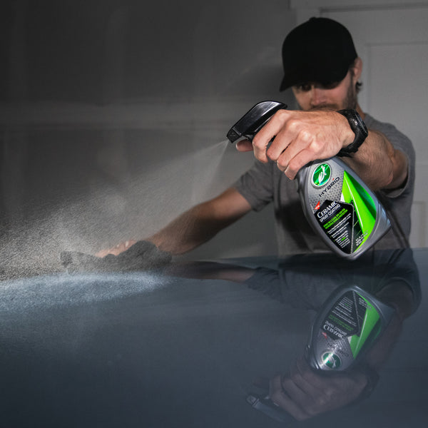 Action shot of HS Ceramic Spray being applied directly to a car panel, showing a gloved hand holding the bottle mid-spray with visible droplets on the surface.