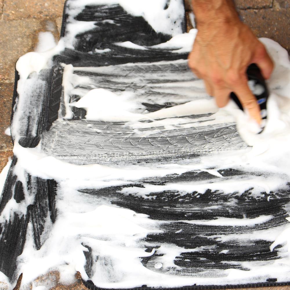Cleaning car mats! A hand scrubs a black vehicle carpet with foaming cleanser, removing grime. Interior detailing maintenance in action.
