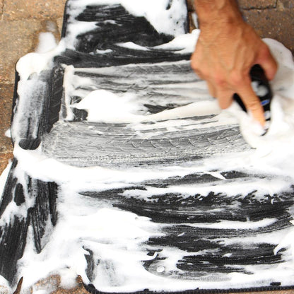 Cleaning car mats! A hand scrubs a black vehicle carpet with foaming cleanser, removing grime. Interior detailing maintenance in action.
