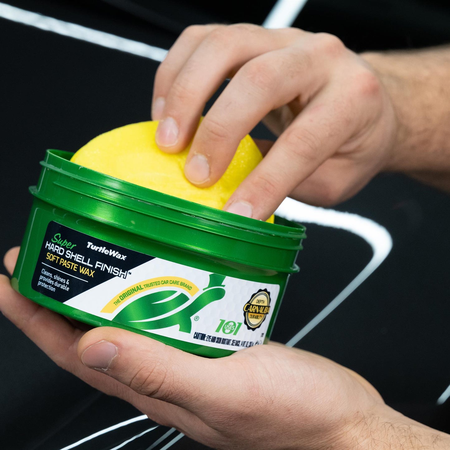 Hands holding Turtle Wax Super Hard Shell Finish with applicator on a car. Green tub, car detailing, trusted car care, durable protection.