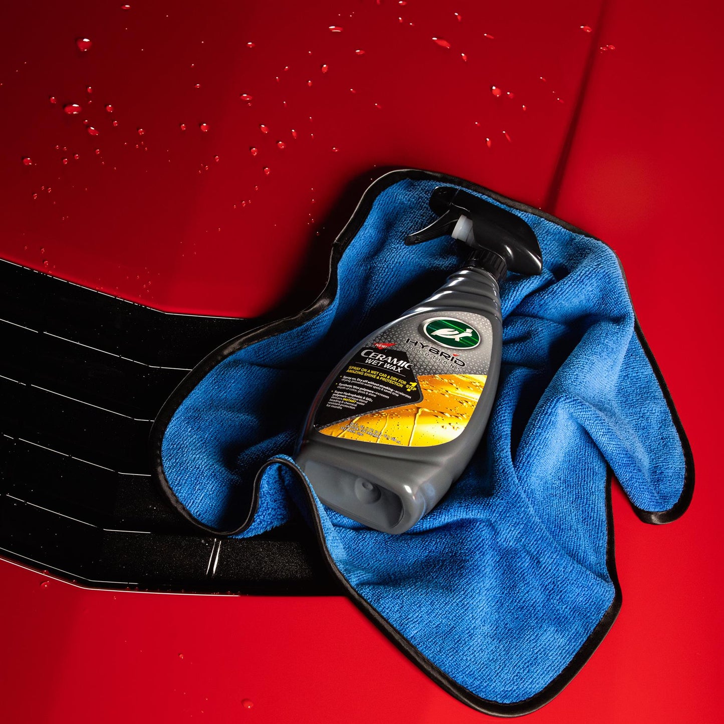 Turtle Wax Hybrid Solutions Ceramic Wet Wax spray sits on a blue microfibre cloth against a shiny red car bonnet background.