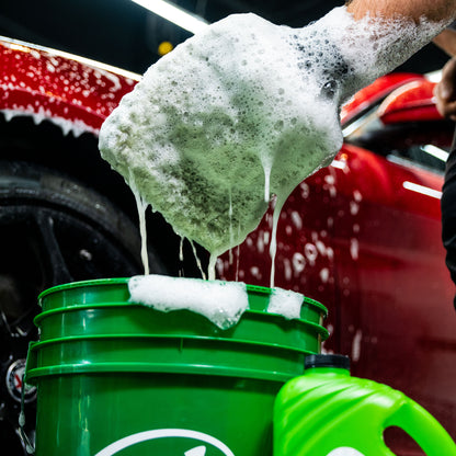 Car care image. A foamy mitt drips suds into a green bucket. Red vehicle nearby.
