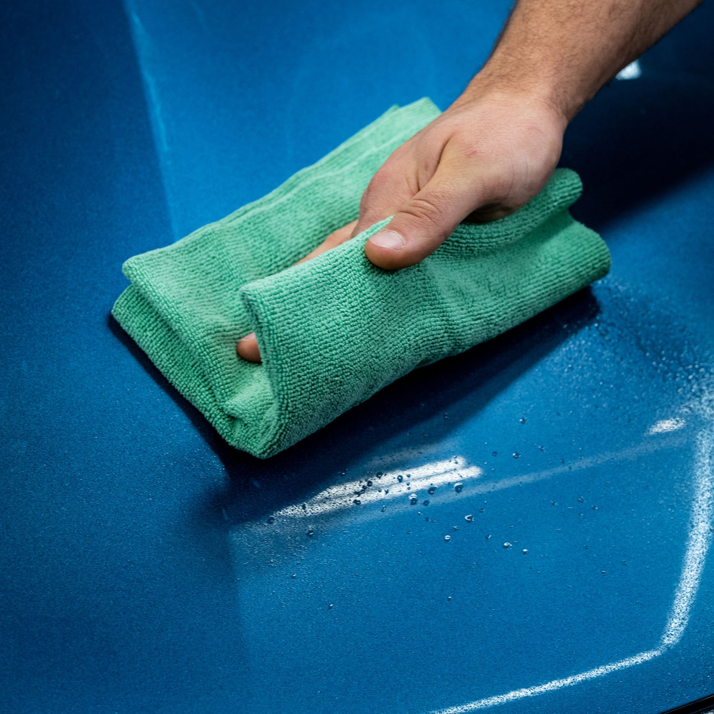Green microfibre cloth buffing blue car paintwork, with slight water beading.