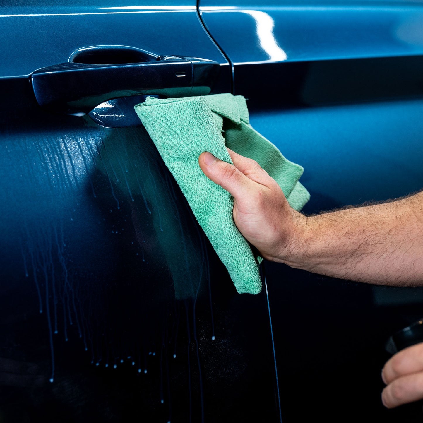 Detailing a blue car. A hand with a green microfibre cloth wipes away liquid streaks by the door handle.