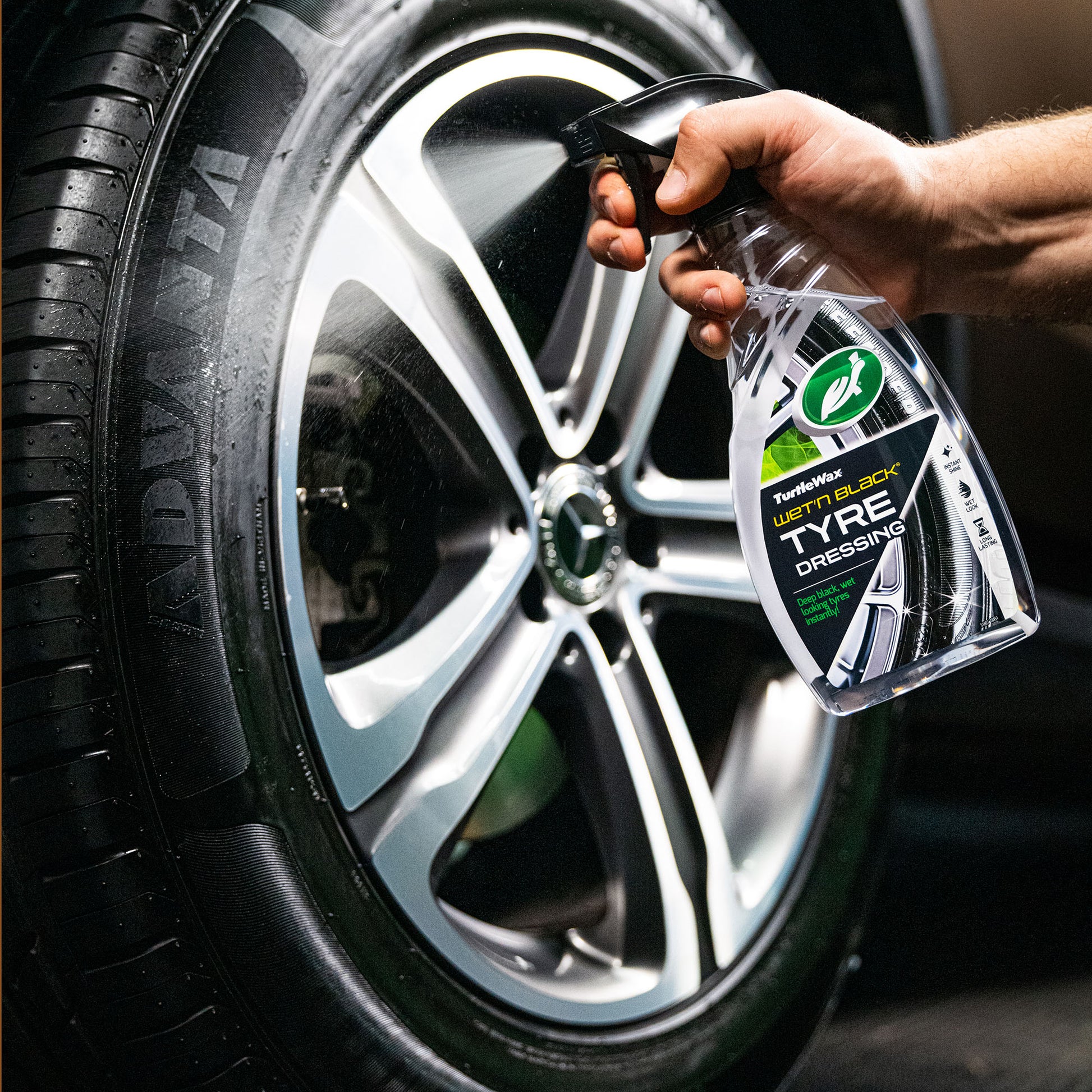 Turtle Wax: close-up of a dark, wet car tyre. The brand's tyre dressing being sprayed onto the rubber by a hand. Modern wheel design.