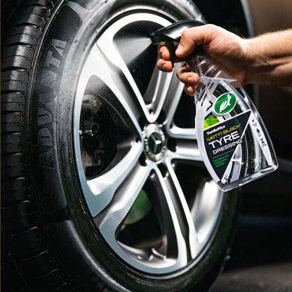 Turtle Wax: close-up of a dark, wet car tyre. The brand's tyre dressing being sprayed onto the rubber by a hand. Modern wheel design.