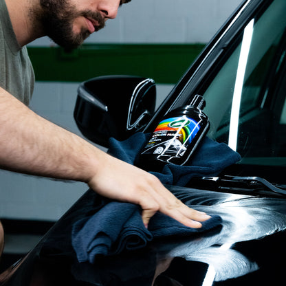 Detailer buffs a dark car using Turtle Wax Color Magic Black Car Polish. Bottle rests on a microfibre cloth.